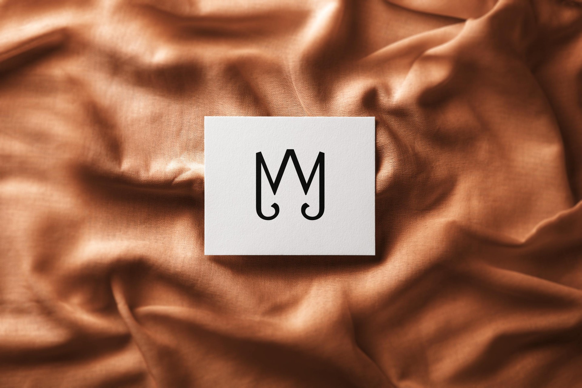 Monogramm-Logo von Martin Joyeux auf weißer Visitenkarte, die auf bronzefarbenem Stoff liegt.